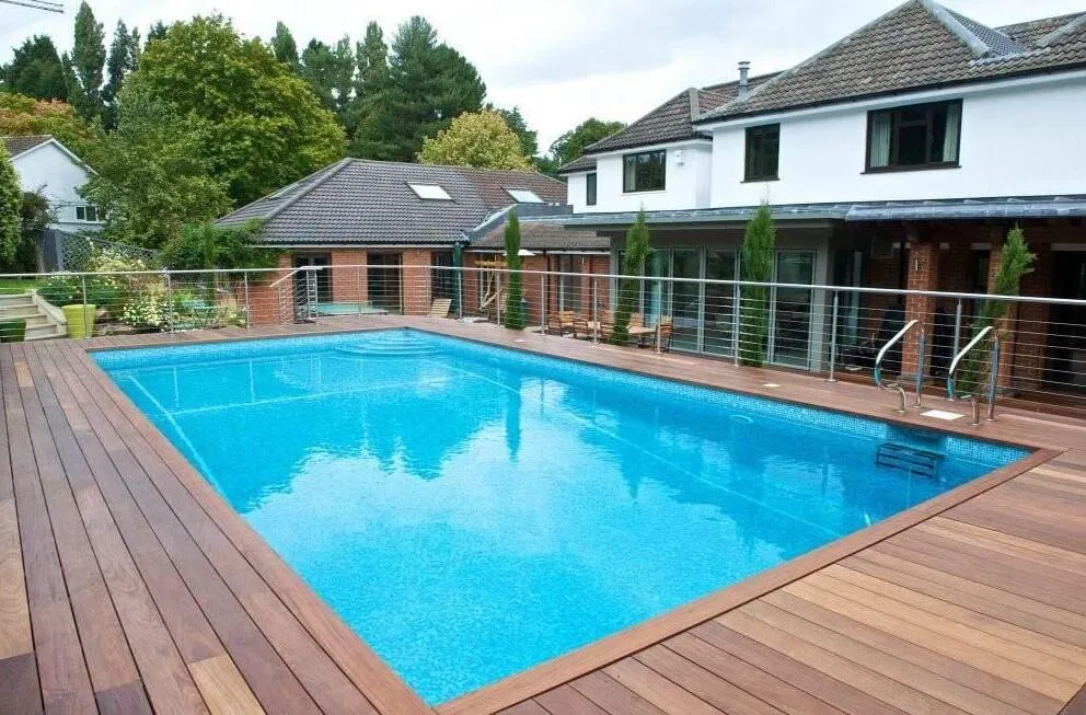 Modern backyard with a large rectangular swimming pool surrounded by a wooden deck and metal railings, attached to a house.