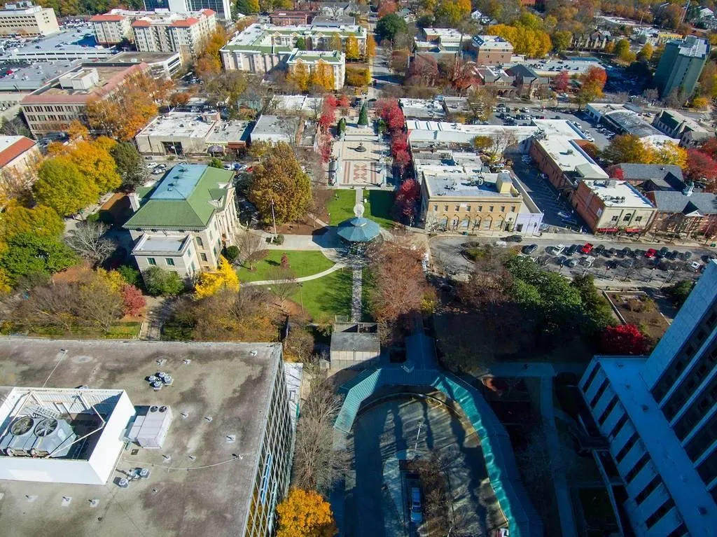 Aerial view of a city park with green trees, surrounding buildings, and colorful autumn foliage in an urban neighborhood