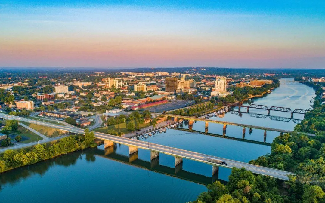 Aerial view of a city skyline with bridges crossing a wide river at sunset, surrounded by green trees and urban buildings