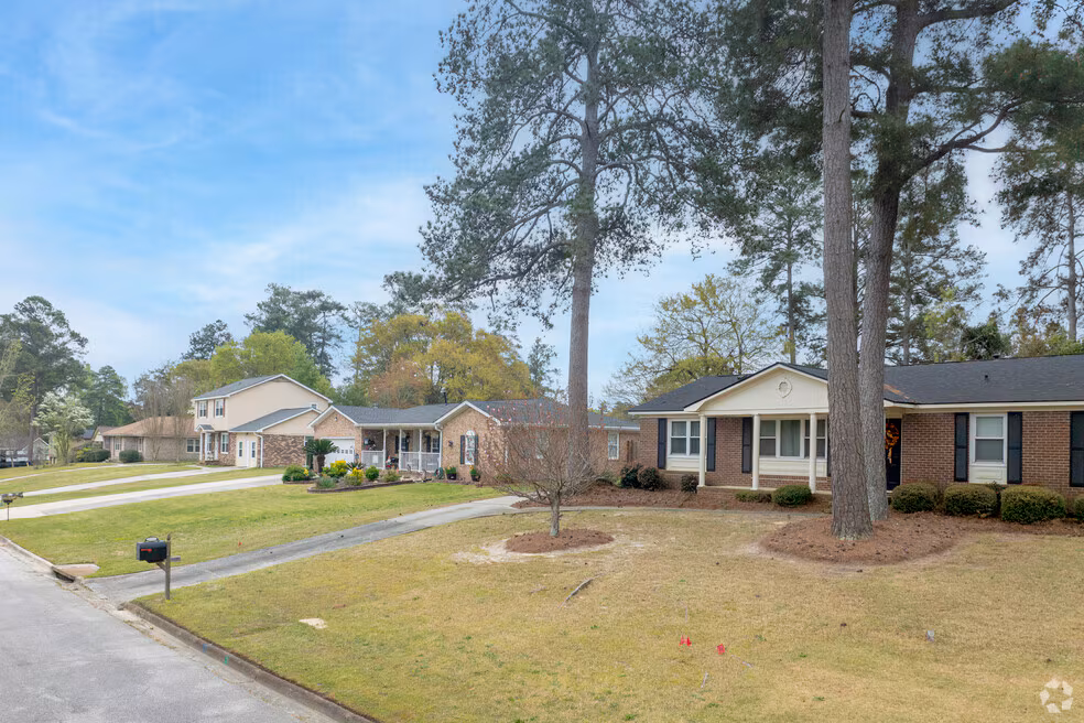 Suburban neighborhood street with single-story brick houses, front lawns, and tall pine trees on a clear day