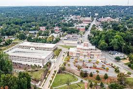 Aerial view of a suburban town with commercial buildings, parking lots, tree-lined streets, and surrounding green landscape