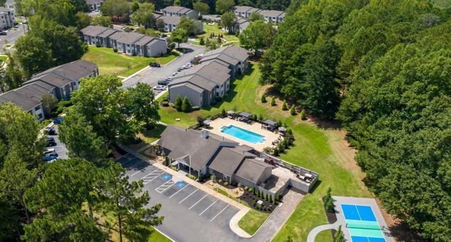 Aerial view of apartment complex with swimming pool, clubhouse, parking lot, and surrounding green trees