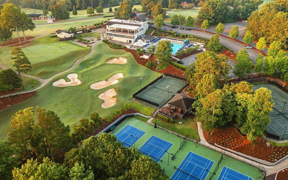 Aerial view of country club with golf course, tennis courts, swimming pool, and clubhouse surrounded by trees