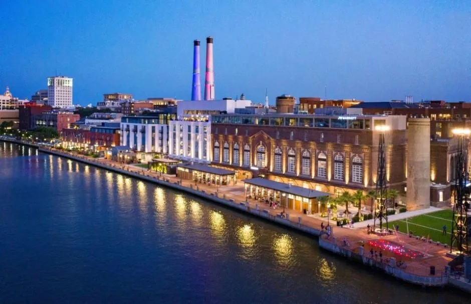 Waterfront cityscape at dusk with historic brick buildings, glowing lights reflecting on the river, and tall smokestacks in the background