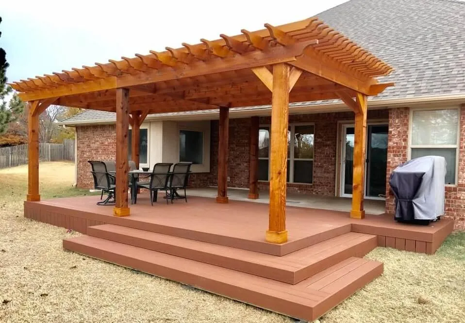Backyard wooden deck with a pergola overhead, outdoor dining set, and grill, attached to a brick house.