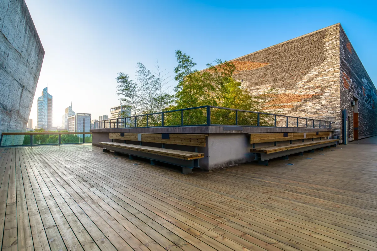 Spacious rooftop wooden deck with built-in benches and greenery in the center, surrounded by tall brick walls and city views.