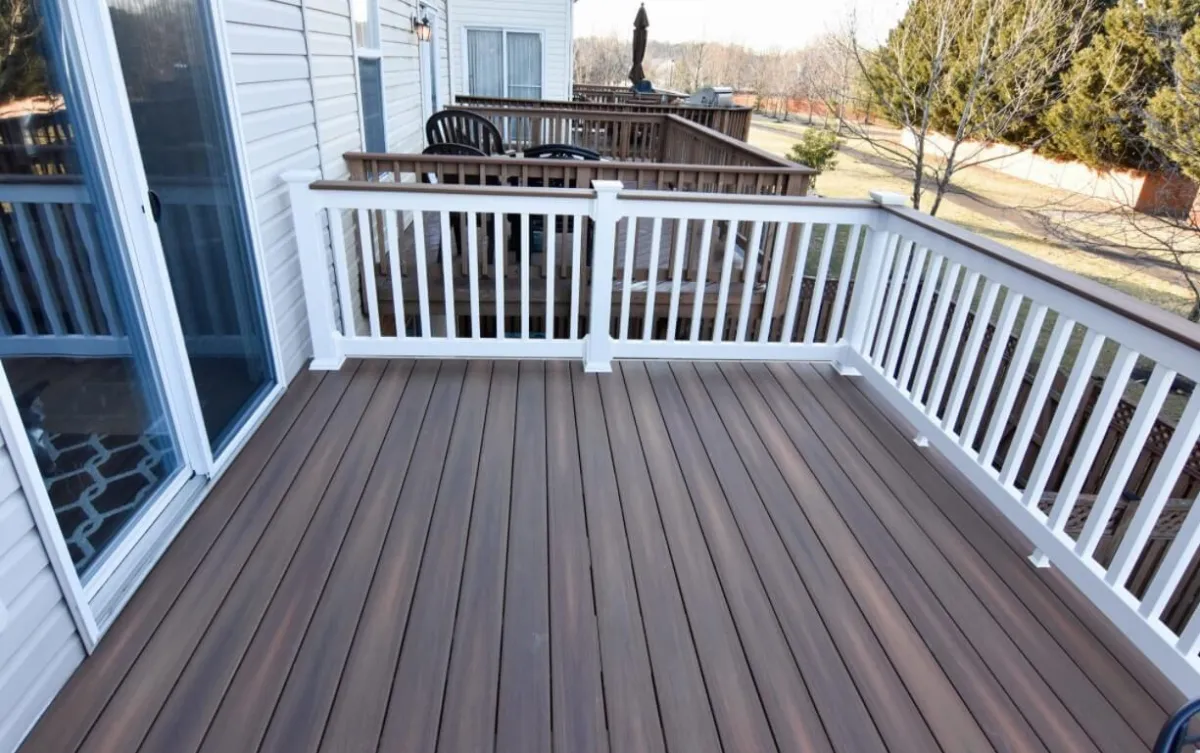 Wooden backyard deck with brown flooring and white railings, attached to a house with sliding glass doors.