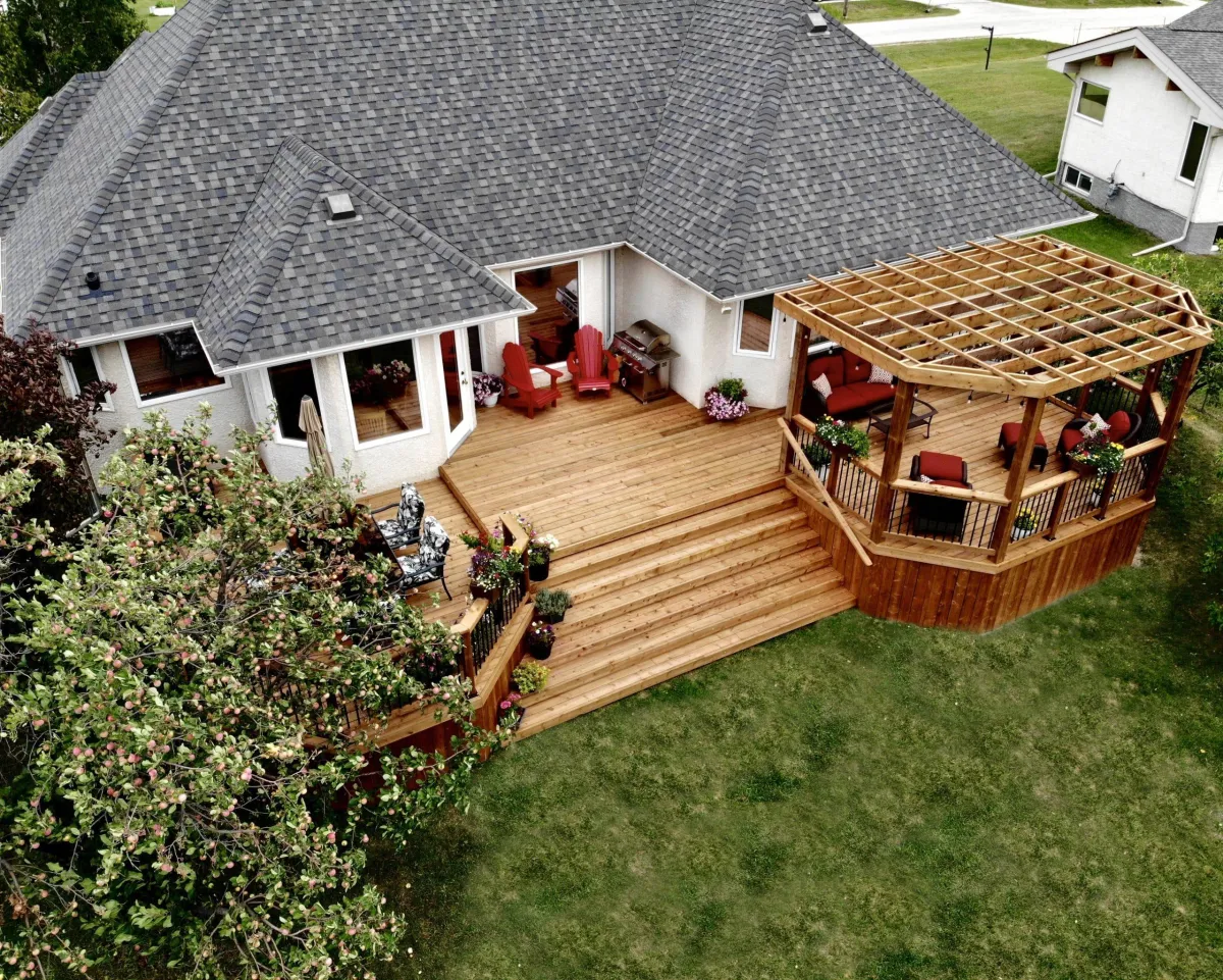 Aerial view of a house with a large wooden backyard deck featuring wide steps, seating areas, and a pergola-covered lounge.