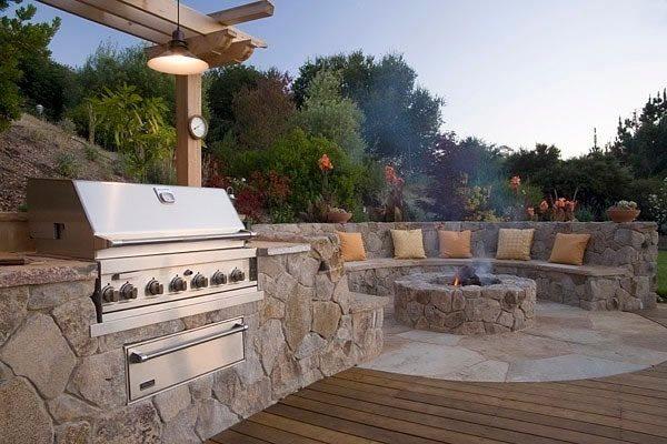 Outdoor patio with built-in stone grill, circular stone fire pit, and curved stone bench seating with cushions.