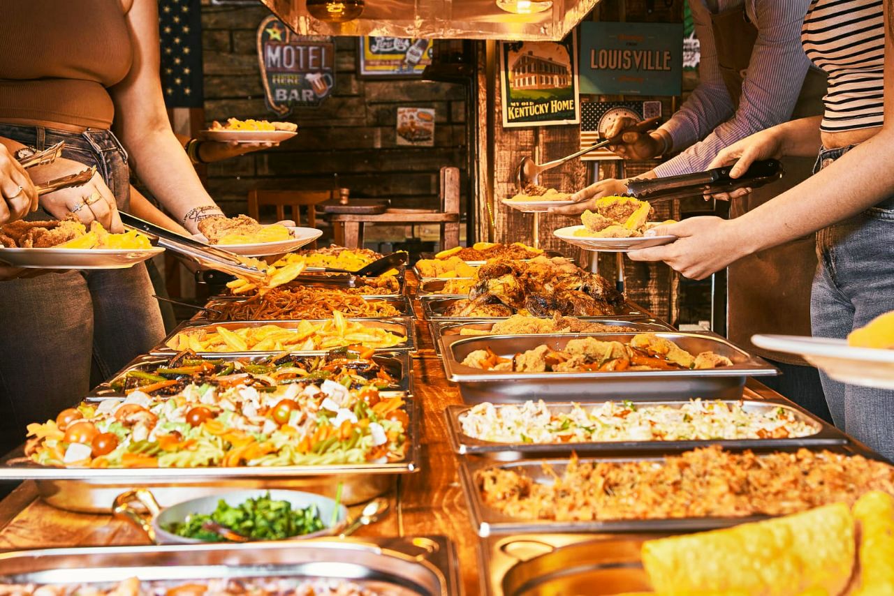 Bright buffet restaurant with a variety of Mediterranean dishes, fresh salads, and fruits on display. Large windows let in natural light, and tables are set for groups, creating a lively, inviting atmosphere.