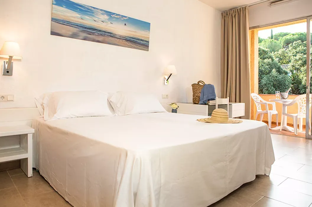 Spacious, sunlit hotel room with two queen beds, crisp white linens, blue accent pillows, and a balcony overlooking the Mediterranean. The room is modern, airy, and designed for team comfort.