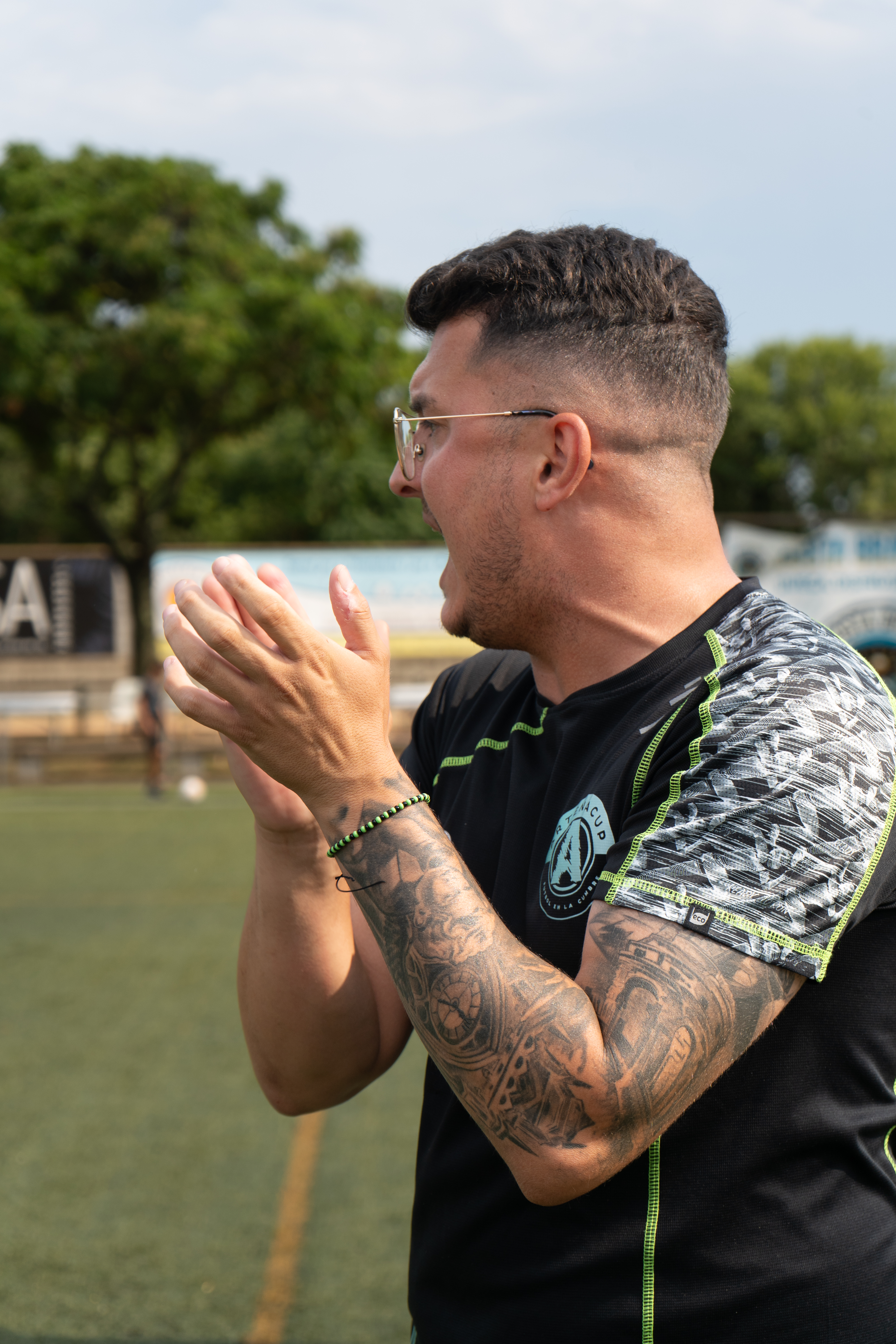 Smiling football coach in a blue polo, holding a clipboard, standing on a sunlit Mediterranean terrace with the sea in the background. The coach appears relaxed and confident, embodying leadership and hospitality.