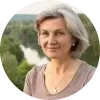 Portrét usmievajúcej sa strednovekej ženy so šedivými vlasmi, oblečenej v hnedosivom tričku, s prírodným pozadím rieky a zelene.