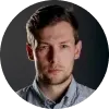 Portrét vážne vyzerajúceho mladého muža s krátkymi hnedými vlasmi, oblečeného v pruhovanej modro-bielej košeli.