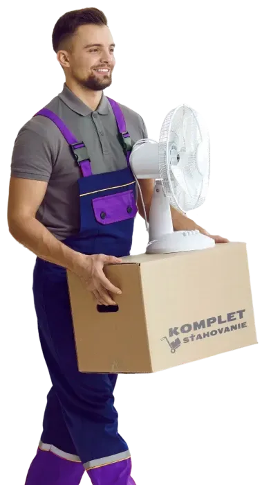 Usmievavý sťahovací pracovník v šedom tričku a fialových montérkach drží kartónovú krabicu označenú s logom Komplet Sťahovanie a biely stolný ventilátor.