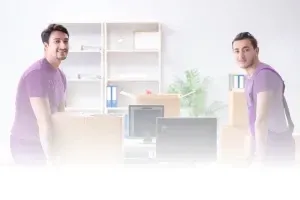 Dvaja muži v fialových tričkách a montérkach stoja v kancelárii obklopení krabicami a nábytkom, pripravení na sťahovanie.