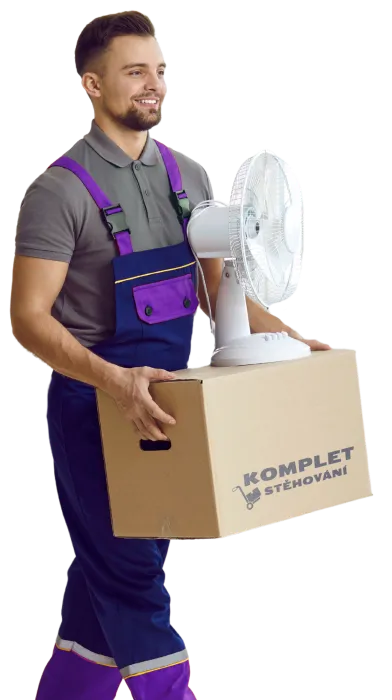 Pracovník týmu "Komplet Stěhování" ve fialových montérkách a šedém tričku, nese kartonovou krabici s logem "Komplet Stěhování" a na ní je postavený stolní ventilátor. Muž se usmívá a kráčí vpřed.