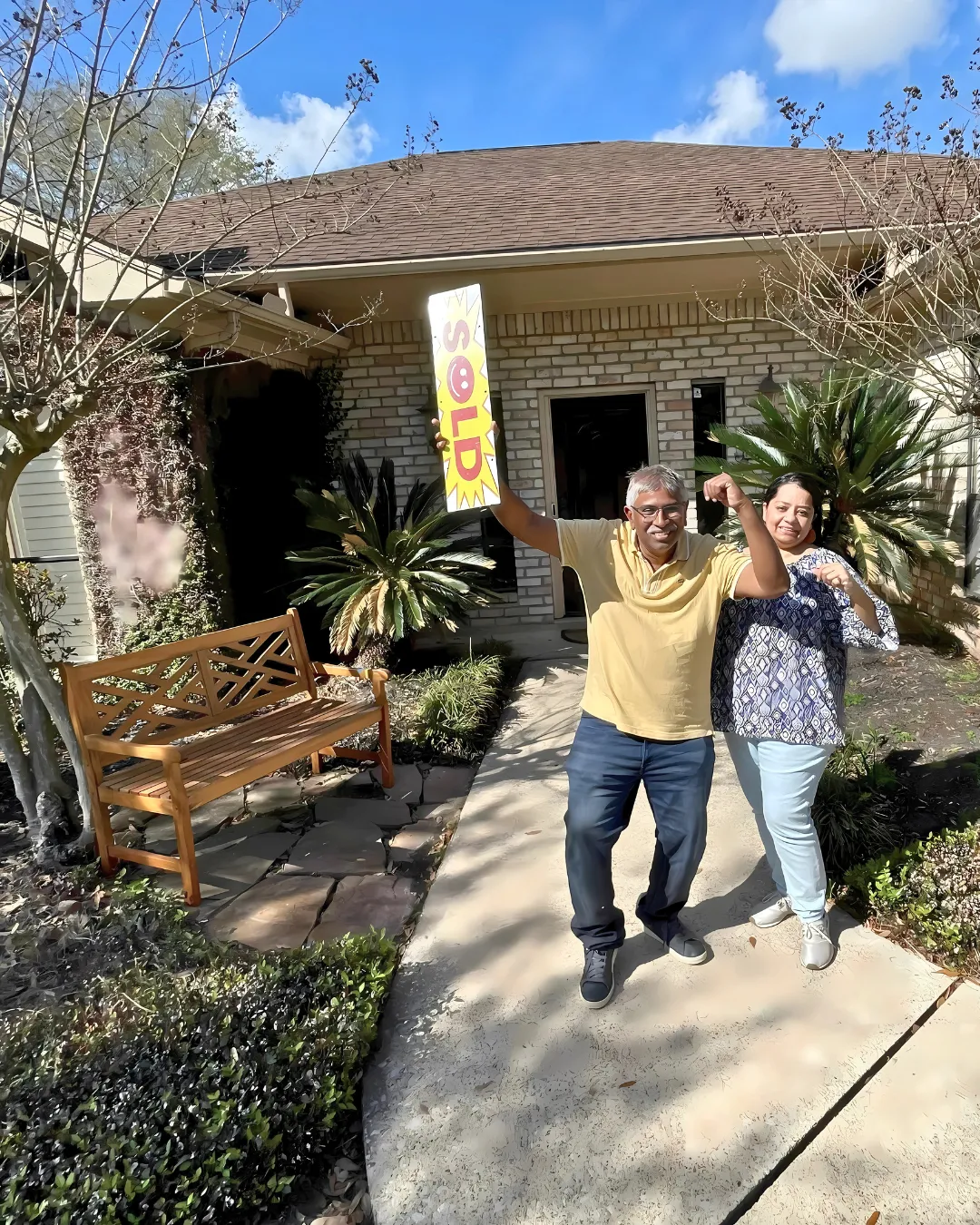 Happy couple celebrating home purchase with "SOLD" sign in front of their new house, surrounded by greenery and a wooden bench.