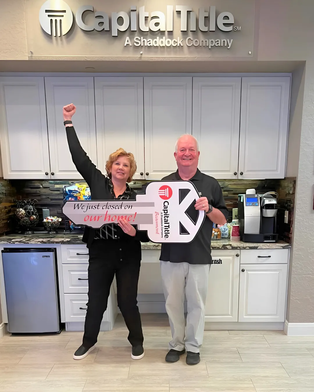 Couple celebrating home closing with oversized key at Capital Title office, showcasing excitement and success in real estate transaction.