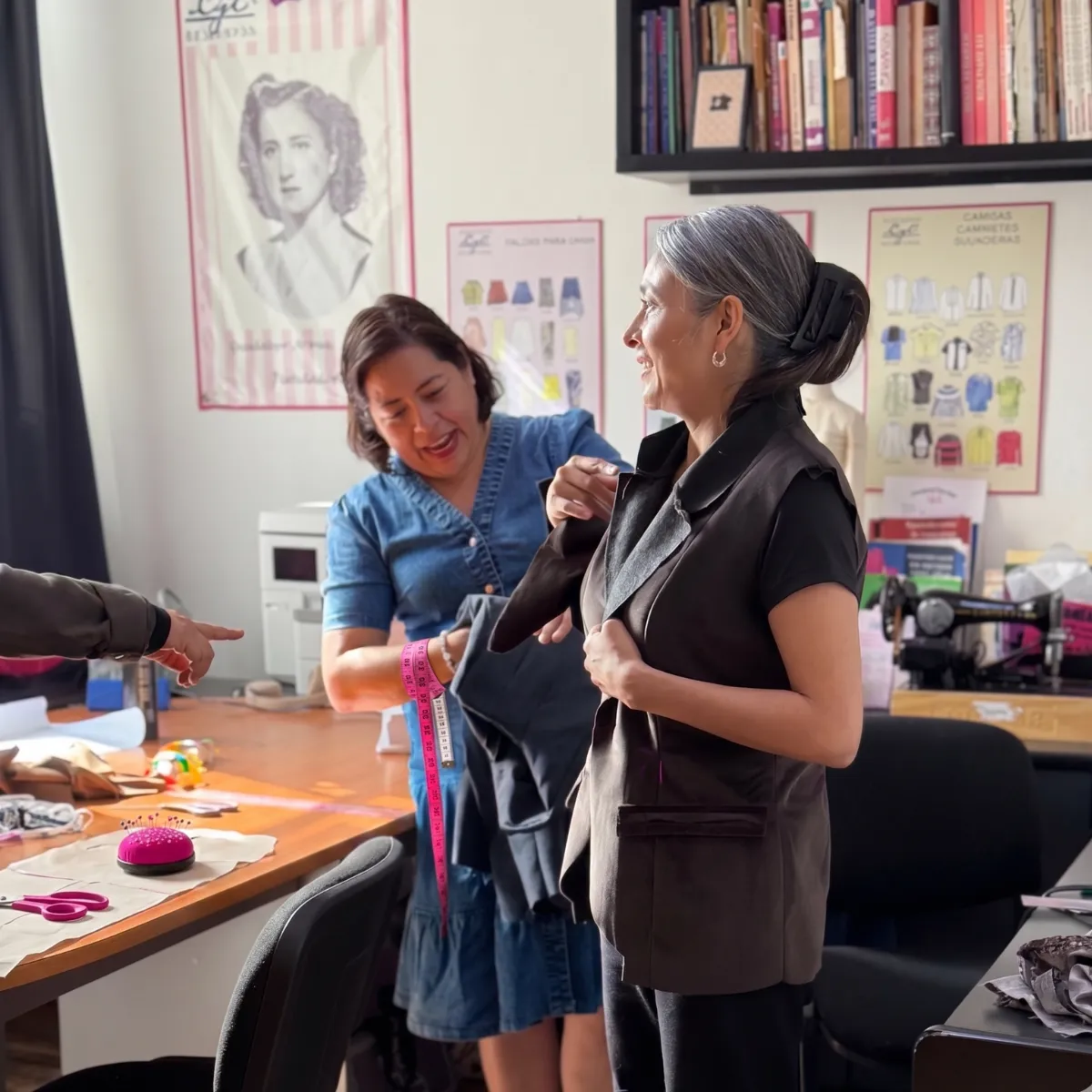 Dos mujeres en una sesión práctica de ajuste de prendas en un taller de costura; al fondo se observa el retrato de la fundadora de Sistema CyC y materiales didácticos, representando la facilidad y tradición del método desde 1935.