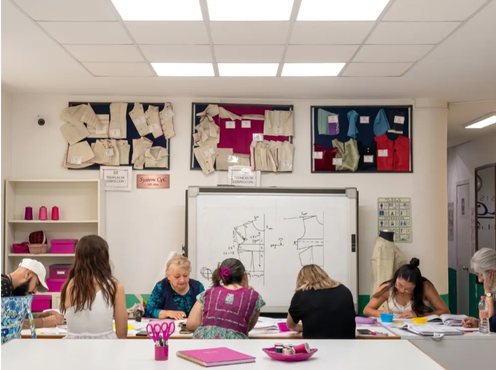 Primer plano de un grupo de estudiantes y una instructora de "Sistema CyC" en una mesa de trabajo, concentrados en aprender técnicas de confección y moldería de prendas sobre papel.