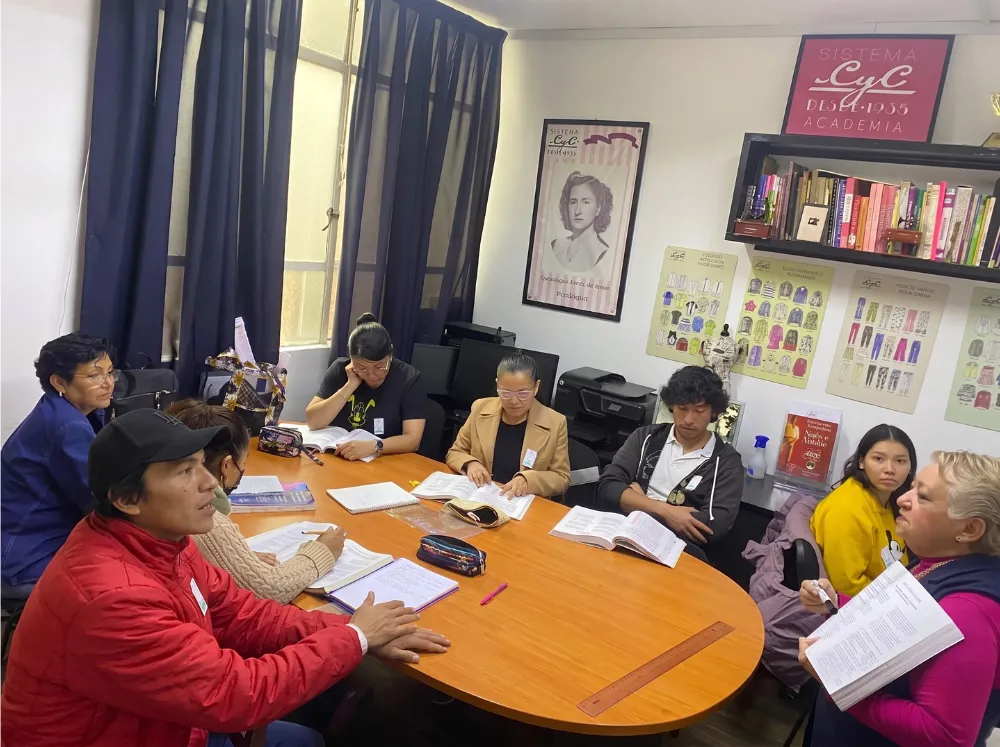 Grupo diverso de estudiantes en una clase presencial del curso de primeras puntadas de Sistema CyC, aprendiendo patronaje y confección de ropa personalizada alrededor de una mesa de estudio con libros didácticos del Sistema CyC