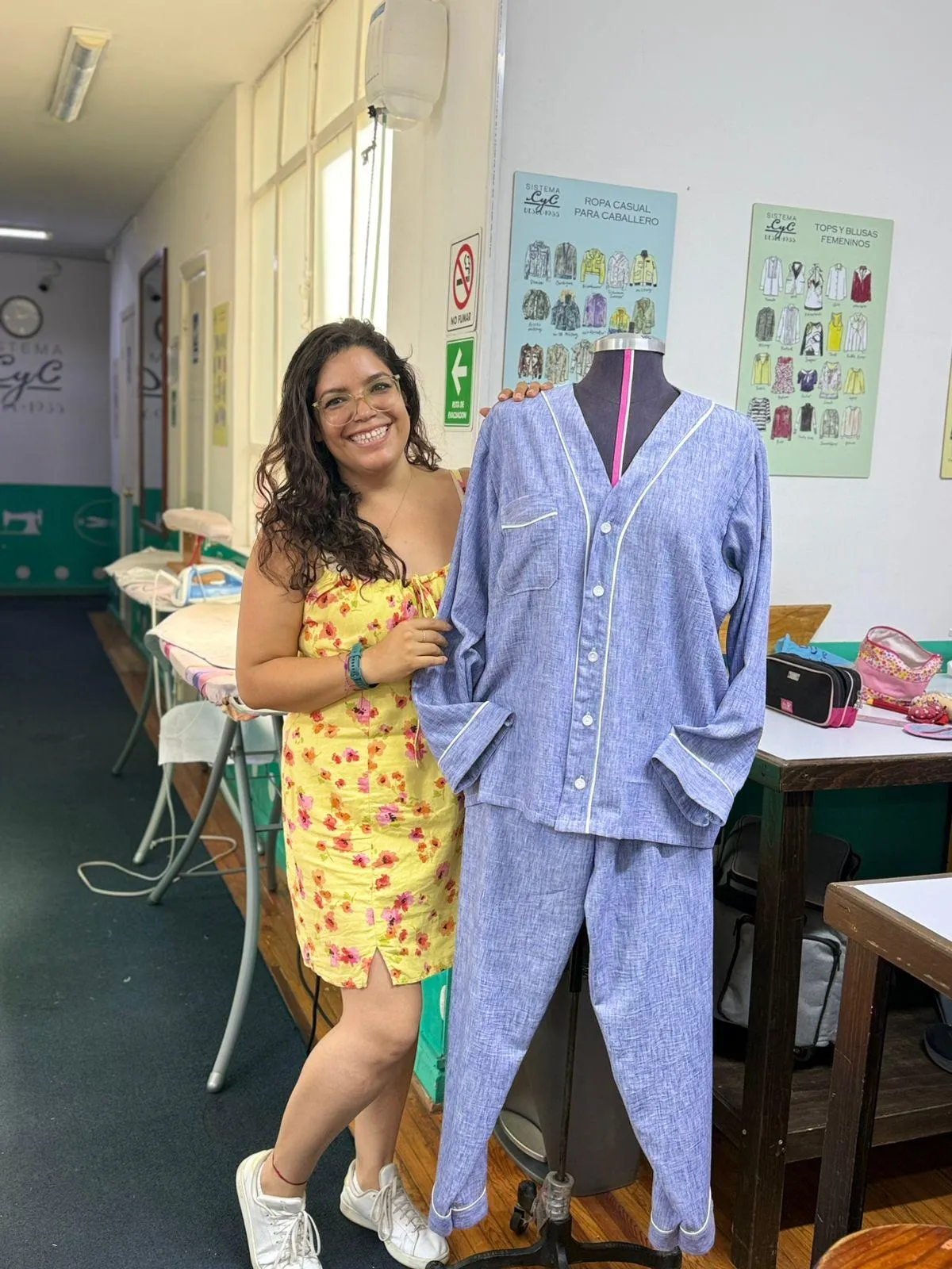 Una estudiante sonriente de la Academia CyC posa con orgullo junto a un maniquí que viste una pijama azul claro con vivos blancos, confeccionada por ella misma. La fotografía está tomada en un taller de costura real donde se aprecia el logotipo de "Sistema CyC desde 1935", burros de planchar y carteles educativos sobre el método de corte y confección. La imagen captura el espíritu de "aprender haciendo" y la satisfacción de alcanzar la autosuficiencia mediante la creación de prendas propias