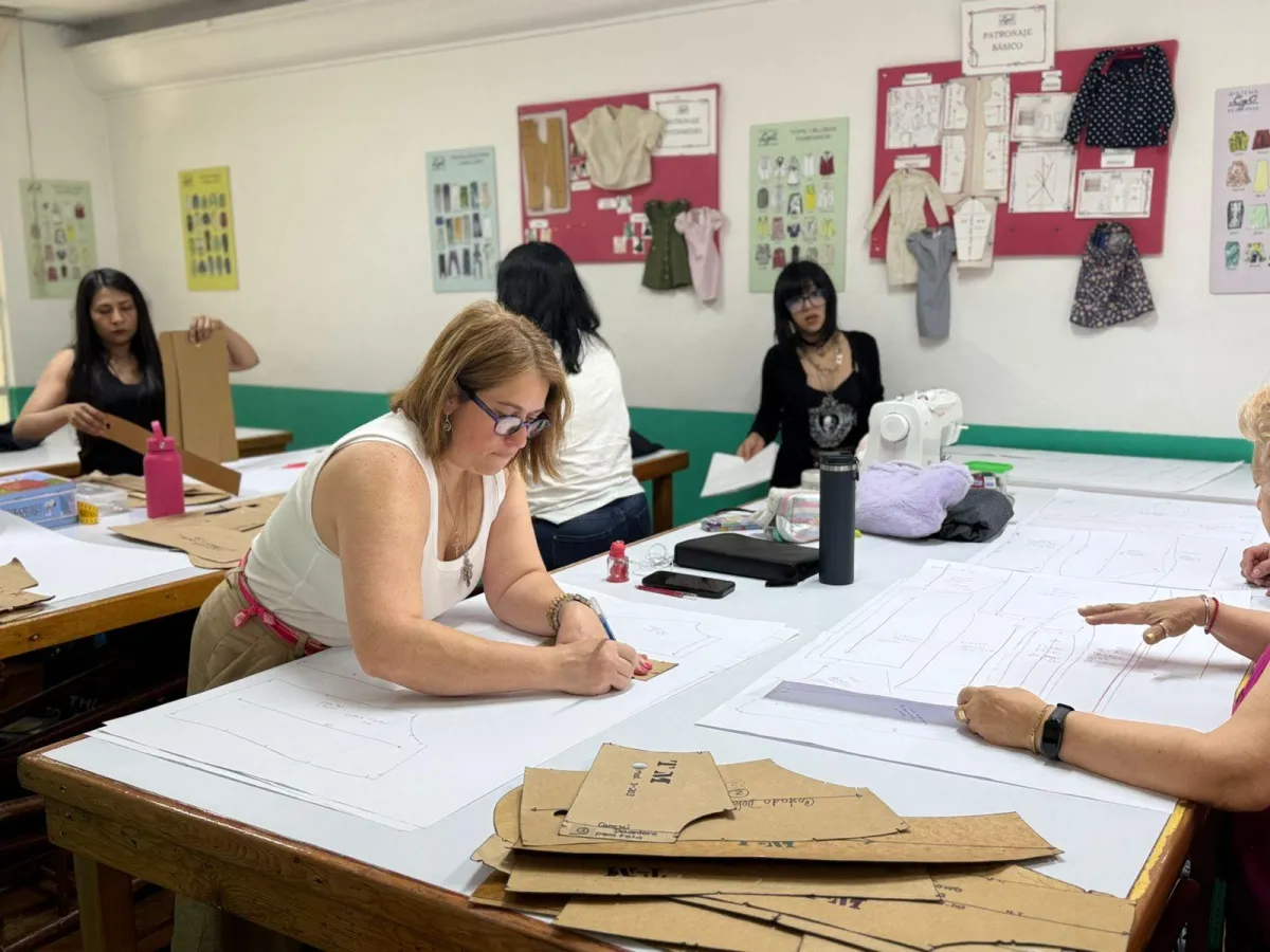 Estudiantes en una clase presencial de la Academia CyC trabajando en el trazo de plantillas básicas y patrones sobre papel en mesas de corte. En un ambiente de "aprender haciendo", una mujer en primer plano utiliza herramientas de precisión para transformar sus diseños, mientras al fondo se observan muros con guías de patronaje básico e intermedio que facilitan un aprendizaje fácil y práctico. La imagen ilustra cómo el Sistema CyC profesionaliza el oficio de corte y confección, empoderando a personas creativas para ser autosuficientes en la creación de sus propias prendas