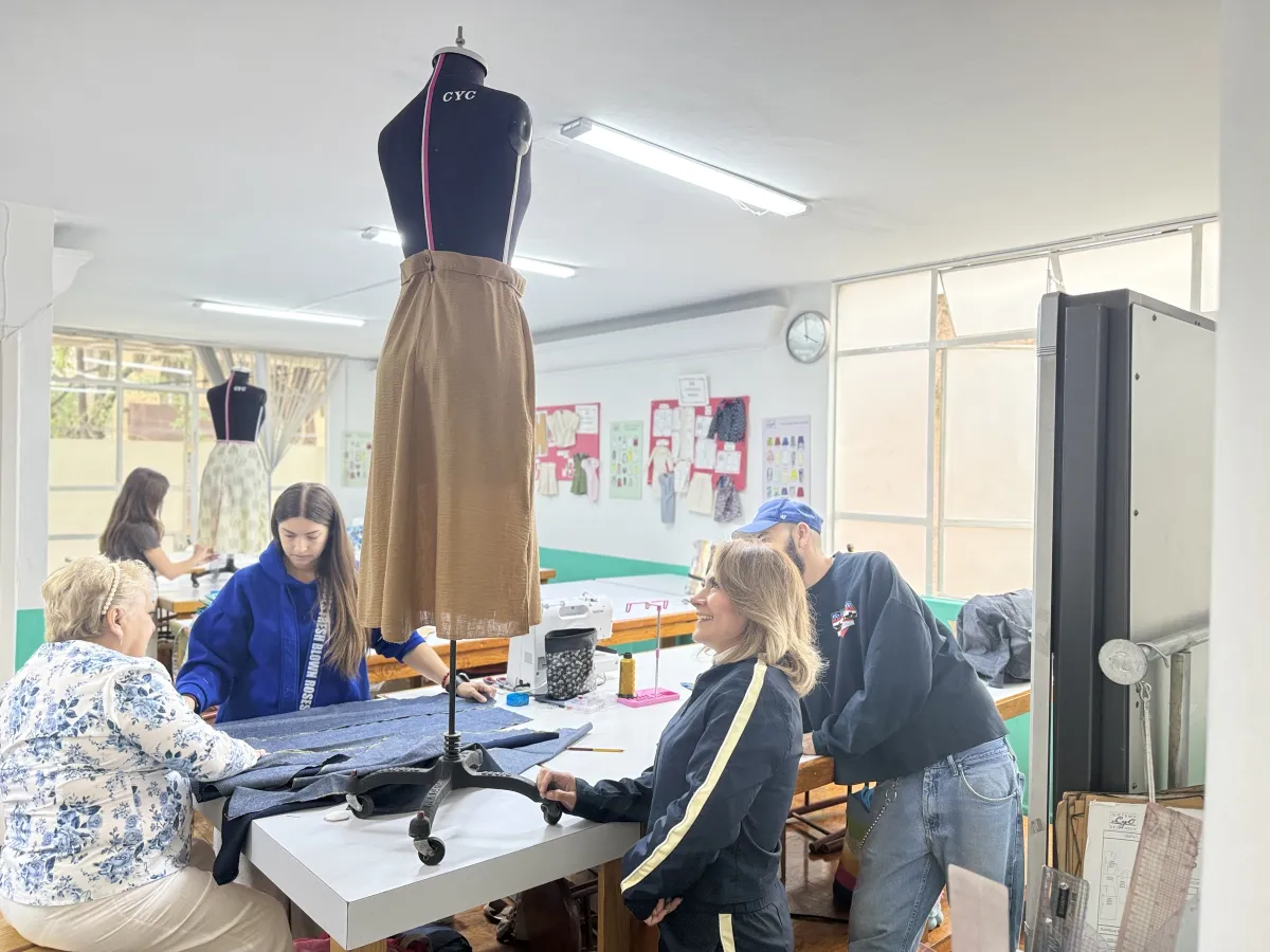 Un grupo diverso de estudiantes e instructores de la Academia CyC colaboran con entusiasmo en un taller luminoso de patronaje y confección. En primer plano, destaca un maniquí de costura con las siglas "CYC" que viste una falda confeccionada en tono café, mientras los alumnos trabajan en equipo analizando textiles sobre una gran mesa de corte con máquinas de coser. Al fondo, se aprecian las aulas equipadas y pizarrones con guías de trazo plano. La imagen transmite un entorno profesional, acogedor y de comunidad, reflejando el orgullo y la atención al detalle de aprender el oficio para ser autosuficientes.