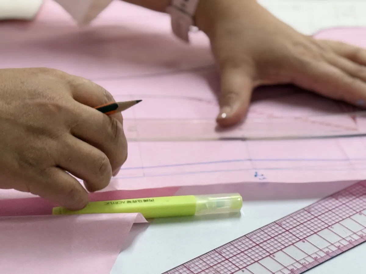 Un grupo diverso de estudiantes e instructores de la Academia CyC colaboran con entusiasmo en un taller luminoso de patronaje y confección. En primer plano, destaca un maniquí de costura con las siglas "CYC" que viste una falda confeccionada en tono café, mientras los alumnos trabajan en equipo analizando textiles sobre una gran mesa de corte con máquinas de coser. Al fondo, se aprecian las aulas equipadas y pizarrones con guías de trazo plano. La imagen transmite un entorno profesional, acogedor y de comunidad, reflejando el orgullo y la atención al detalle de aprender el oficio para ser autosuficientes.