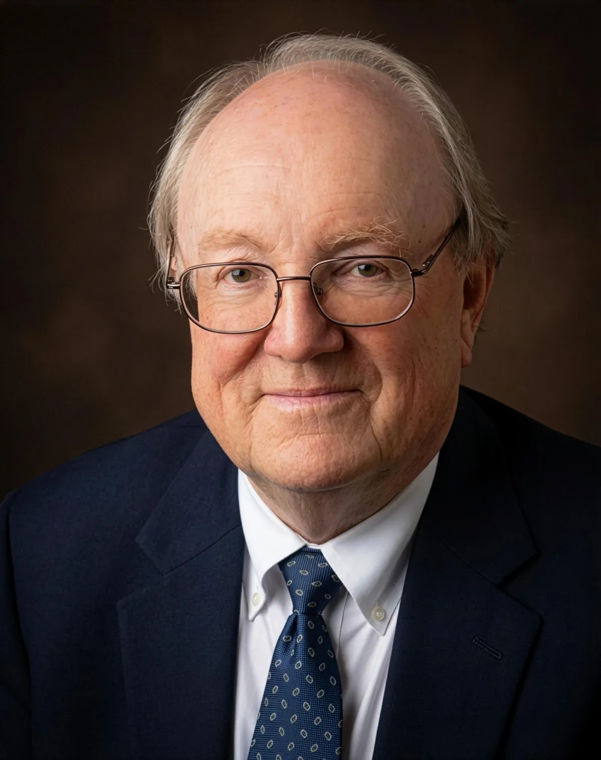 Alt text: Formal portrait of an older man with light hair and glasses, wearing a dark suit, white shirt, and patterned tie, facing the camera against a dark background.