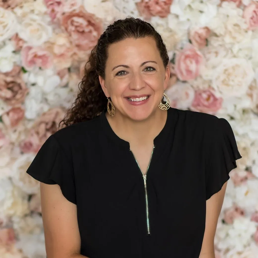 Laurie Walker of LaFaye Photography. Headshot of a woman wearing a black blouse with a zipper detail, long back naturally curly hair, hold hope earings, with white and pink floral background