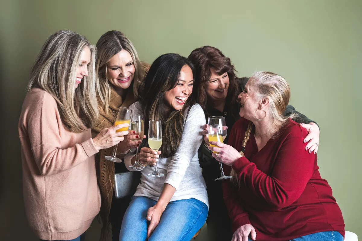 5 ladies together laughing, celebrating their friendship, with wine and champagne having a girls night together,.olive green background, .