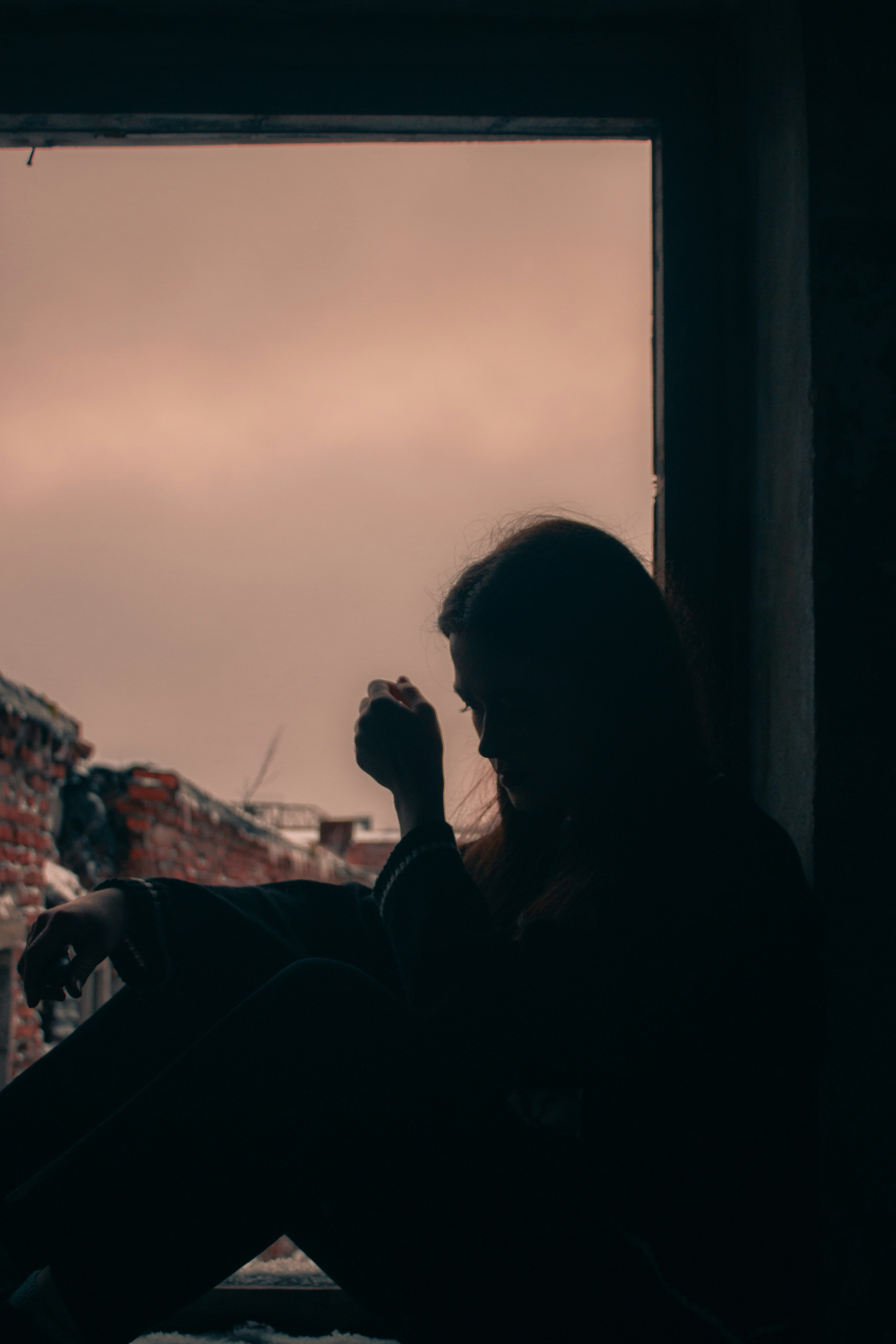 Silhouette of a woman with long hair sitting alone in a darkened doorway against a dusky pink sky, her hand raised thoughtfully to her face, conveying quiet exhaustion and the unseen weight of invisible labor.