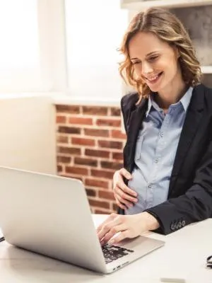 A pregnant woman sits in bed looking at her laptop.