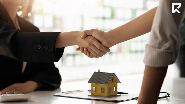 Real estate agent and client shaking hands across a desk with a model house in the foreground, symbolizing a successful property transaction, agreement, or listing appointment. Bright office setting with a professional and welcoming atmosphere.