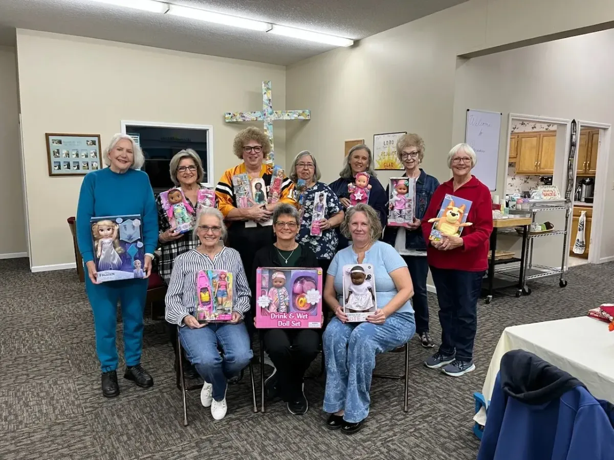 Group of smiling women holding dolls in a church room with a cross on the wall.