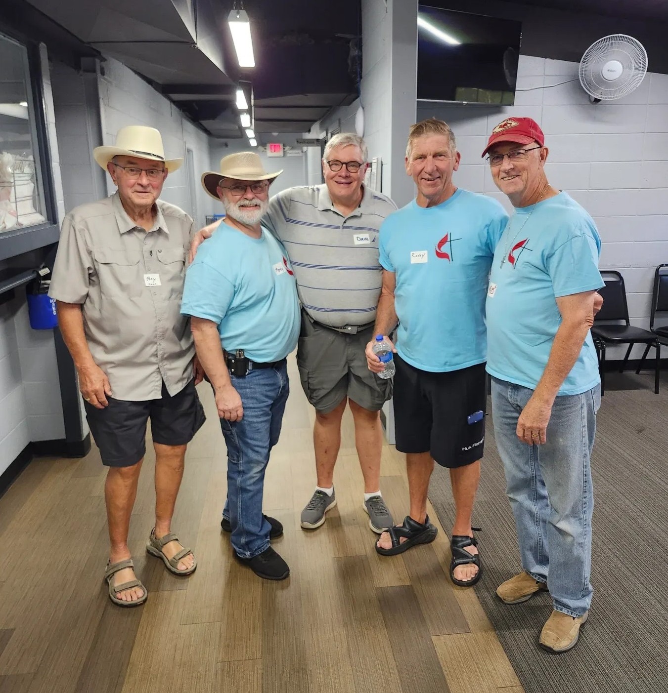 Five men standing indoors, some wearing hats and light blue shirts with name tags.