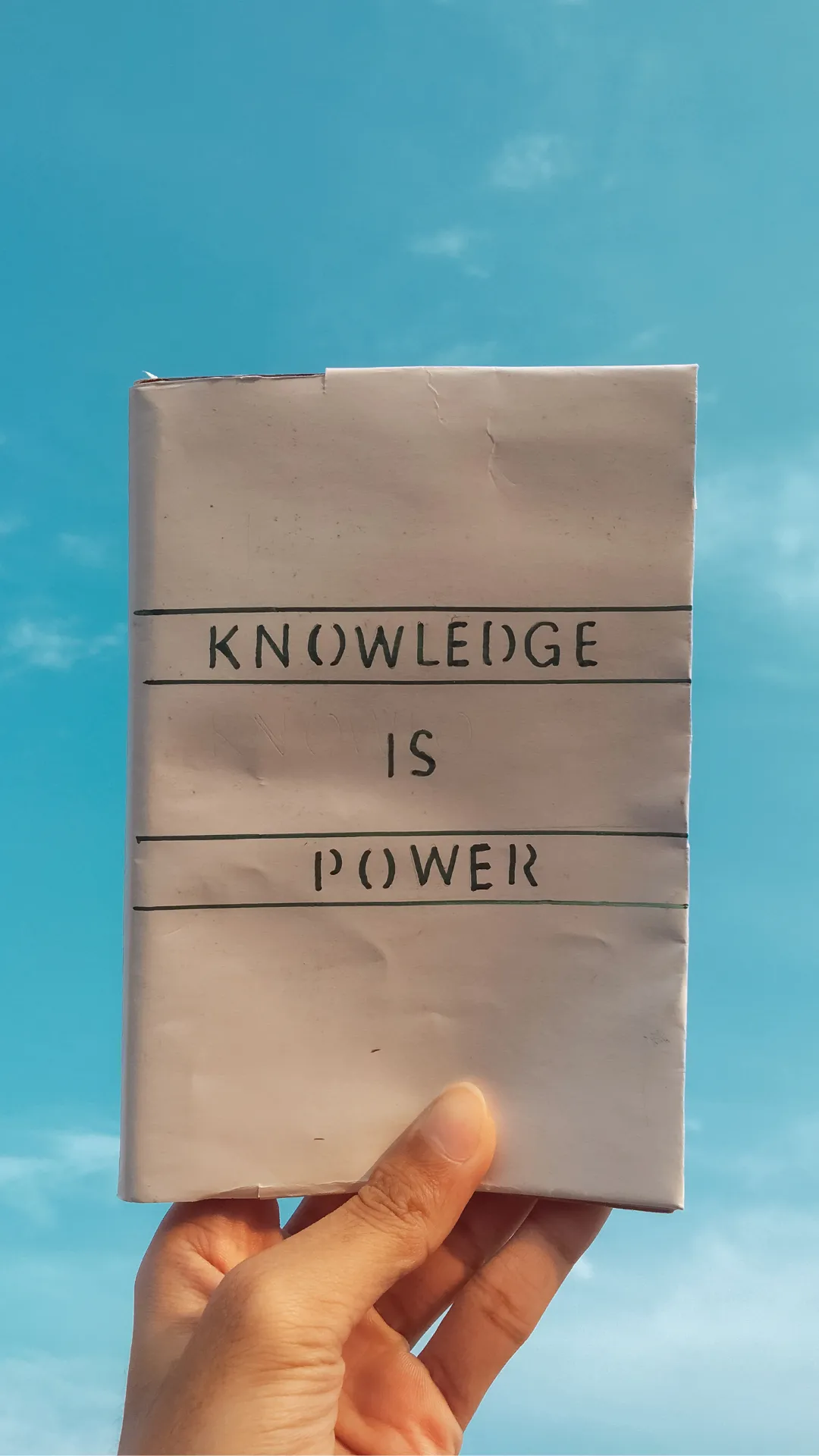 A hand holding up a book against a blue sky, with the words "KNOWLEDGE IS POWER" handwritten on the cover.