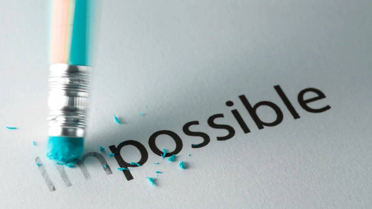 A pencil eraser rubbing out the "im" in "impossible" to reveal the word "possible," with blue eraser shavings on the paper.