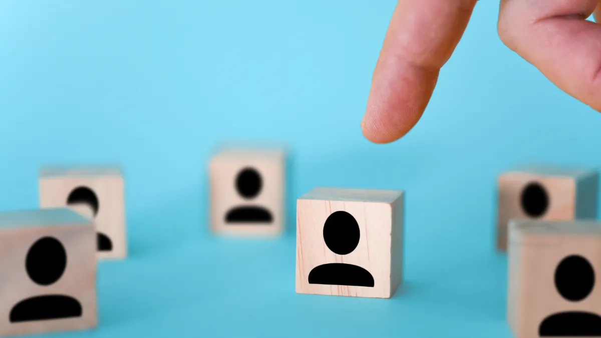A hand pointing towards a wooden cube with a person icon, surrounded by similar cubes, symbolising selection or inclusion.