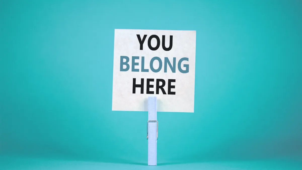 A white card with the words "YOU BELONG HERE" clipped to a blue peg, set against a turquoise background.