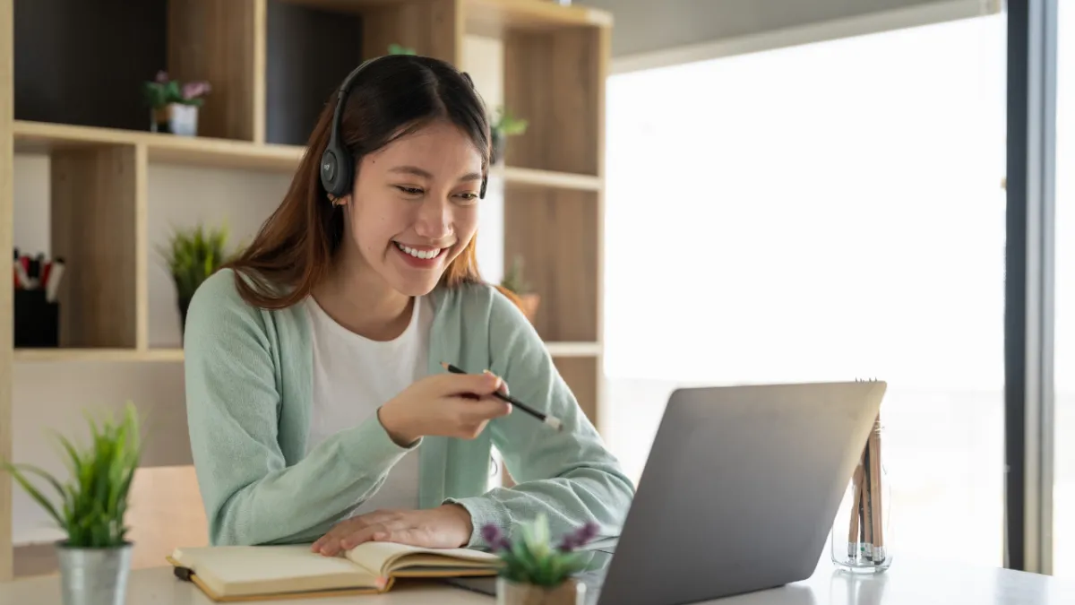 Young woman wearing headphones smiling during an online video call, taking notes in a notebook, symbolising virtual training, coaching or mentoring.