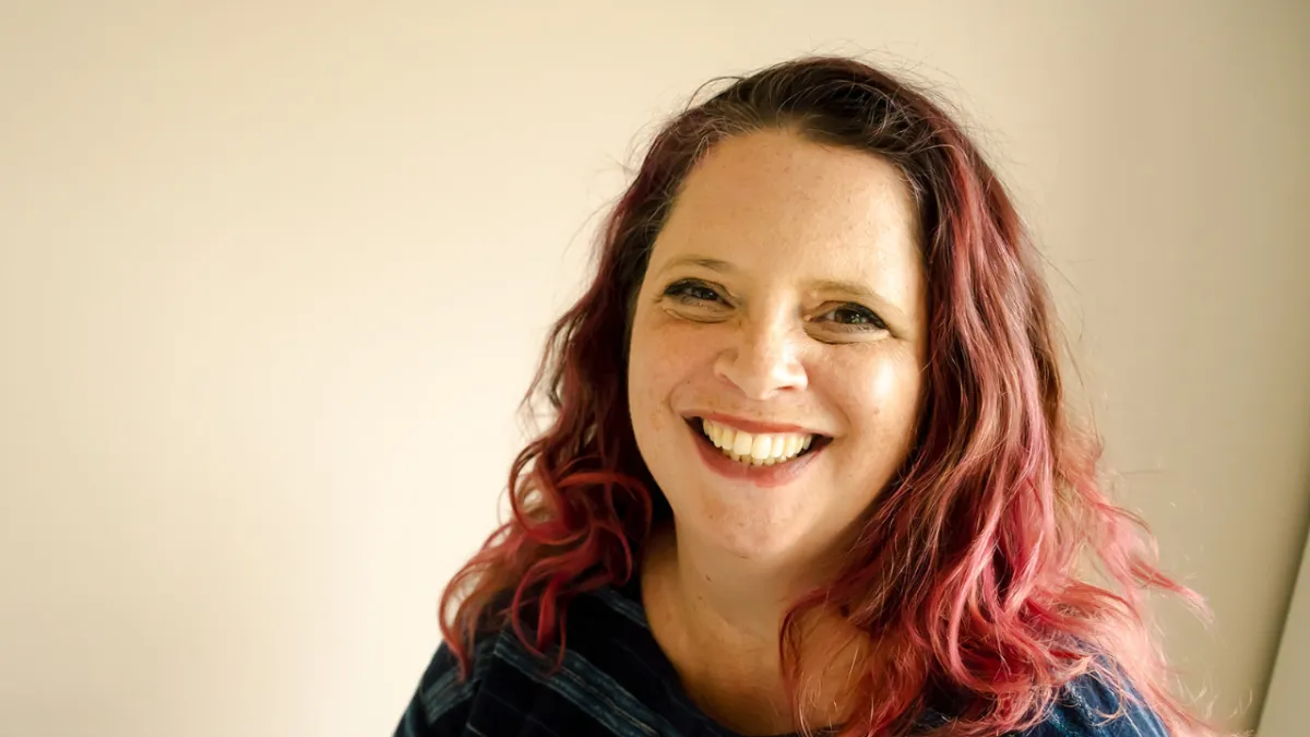 Friendly woman with pink-tinted curly hair smiling warmly against a plain background, representing authentic leadership and wellbeing.