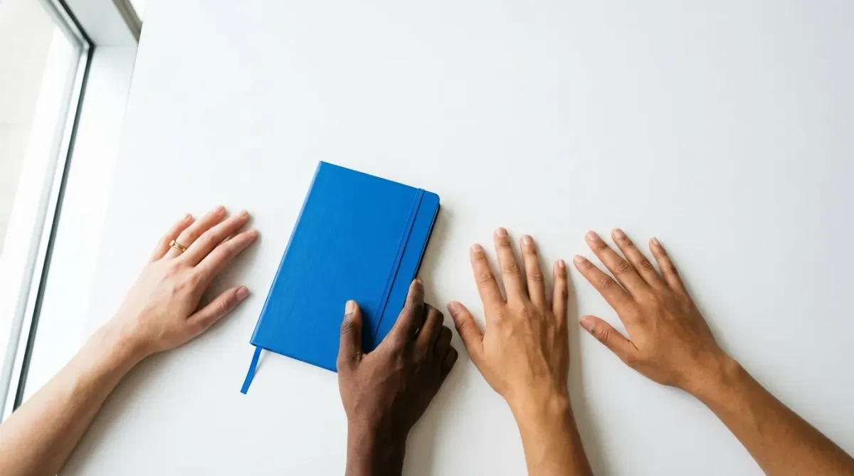 A high-angle, minimalist photo of diverse hands resting on a white table with a vibrant blue notebook, representing collaboration and unity.
