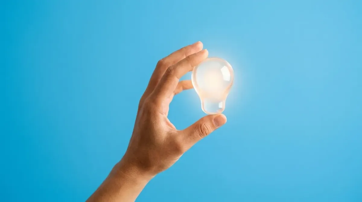 A minimalist photo of a hand holding a glowing light-bulb shape on a bright blue background.
