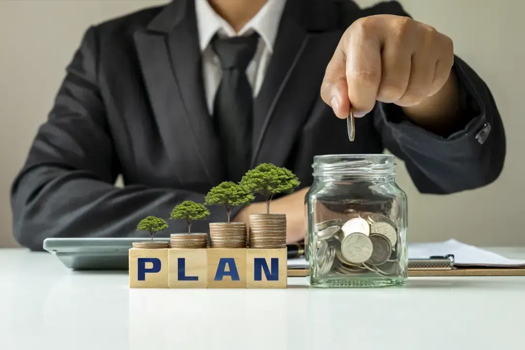 Man tossing a coin into a jar full of coins beside a toy labeled 'PLAN'