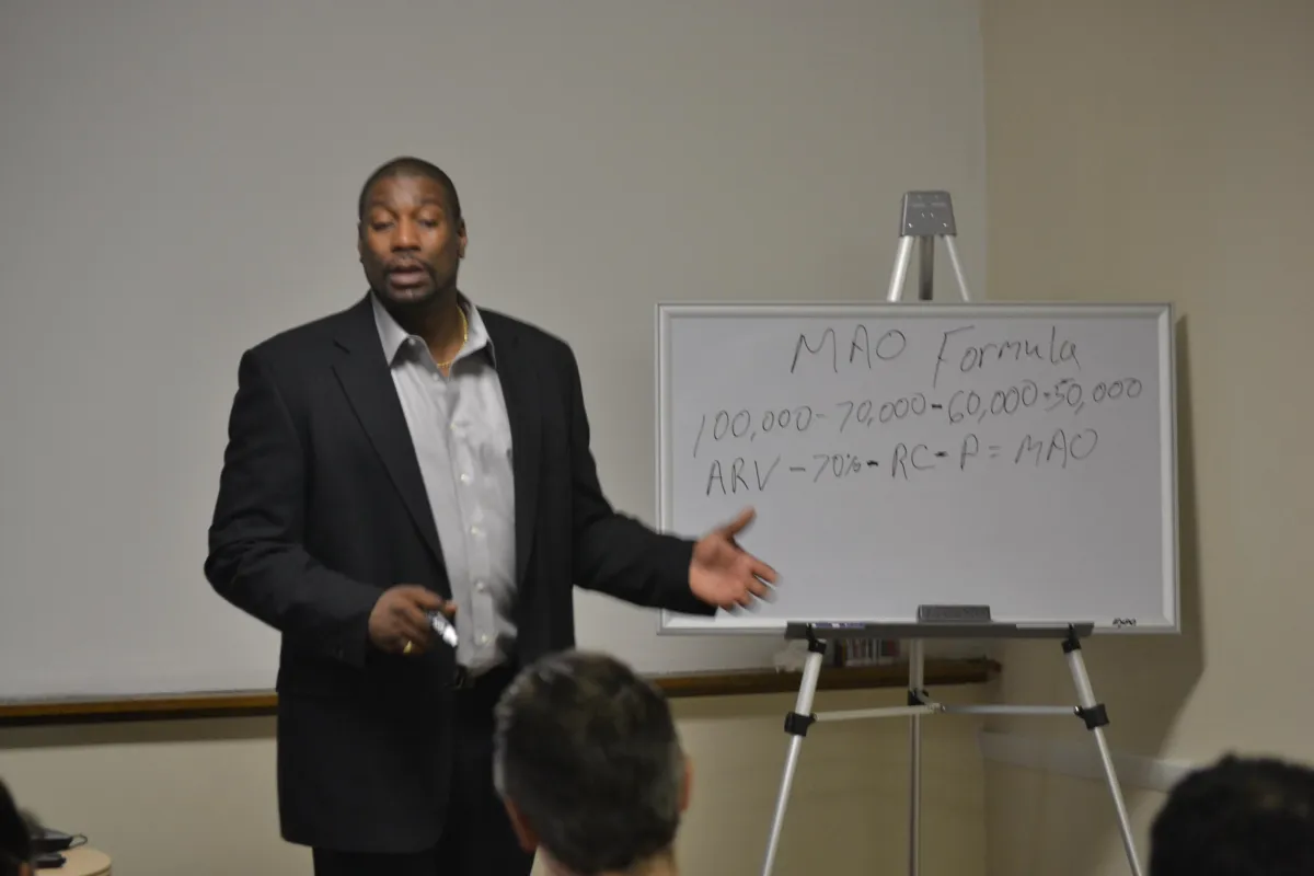 Terence Young teaching a live commercial real estate workshop, standing beside a whiteboard with the MAO Formula (Maximum Allowable Offer) for deal analysis written on it. Demonstrating hands-on training in financial deal structuring.