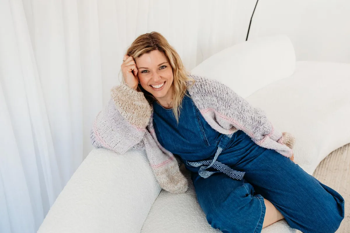 i am Laura chronic illness business, mindset and marketing coach sat on white sofa wearing denim jumpsuit and knitted purple and pink cardigan, smiling at the camera, head leaning on hand