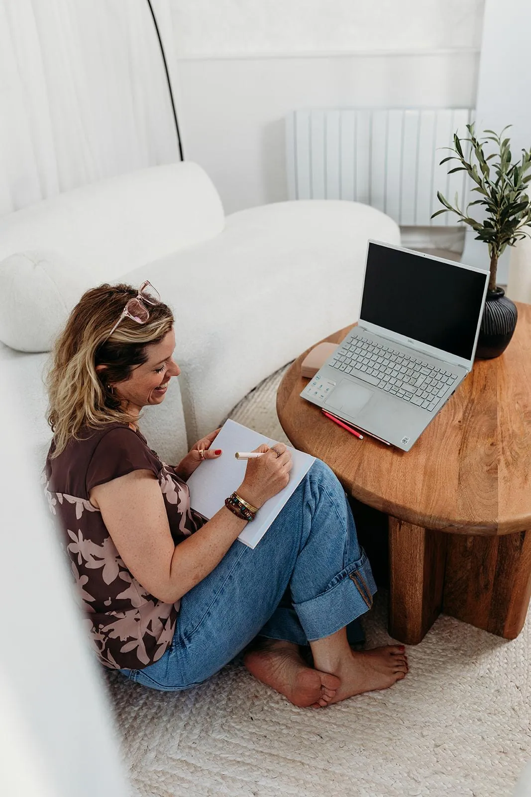 i am Laura chronic illness mums business, marketing and business coach, sitting on floor, looking away writing in note book with laptop in front on table.